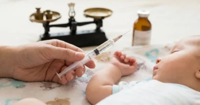Dłoń trzymająca strzykawkę obok śpiącego niemowlęcia, w tle waga szalkowa i butelka z lekiem.
