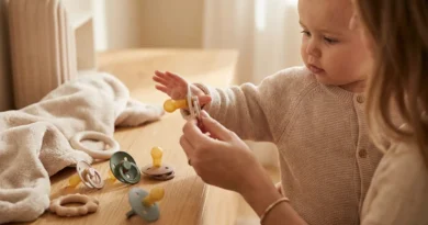 Mama podaje niemowlęciu smoczek; na stole leży kilka innych modeli o różnych kształtach i kolorach.