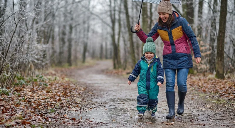 Uśmiechnięta mama i dziecko skaczące w kałuży podczas spaceru w chłodny, deszczowy dzień.