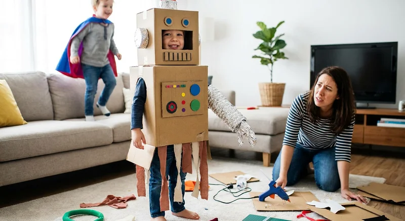 Zmartwiona kobieta z pistoletem na klej obok dziecka w prowizorycznym stroju robota z kartonów.