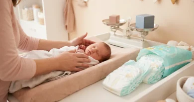 Noworodek na przewijaku obok pieluszek jednorazowych, delikatnie dotykany przez mamę.